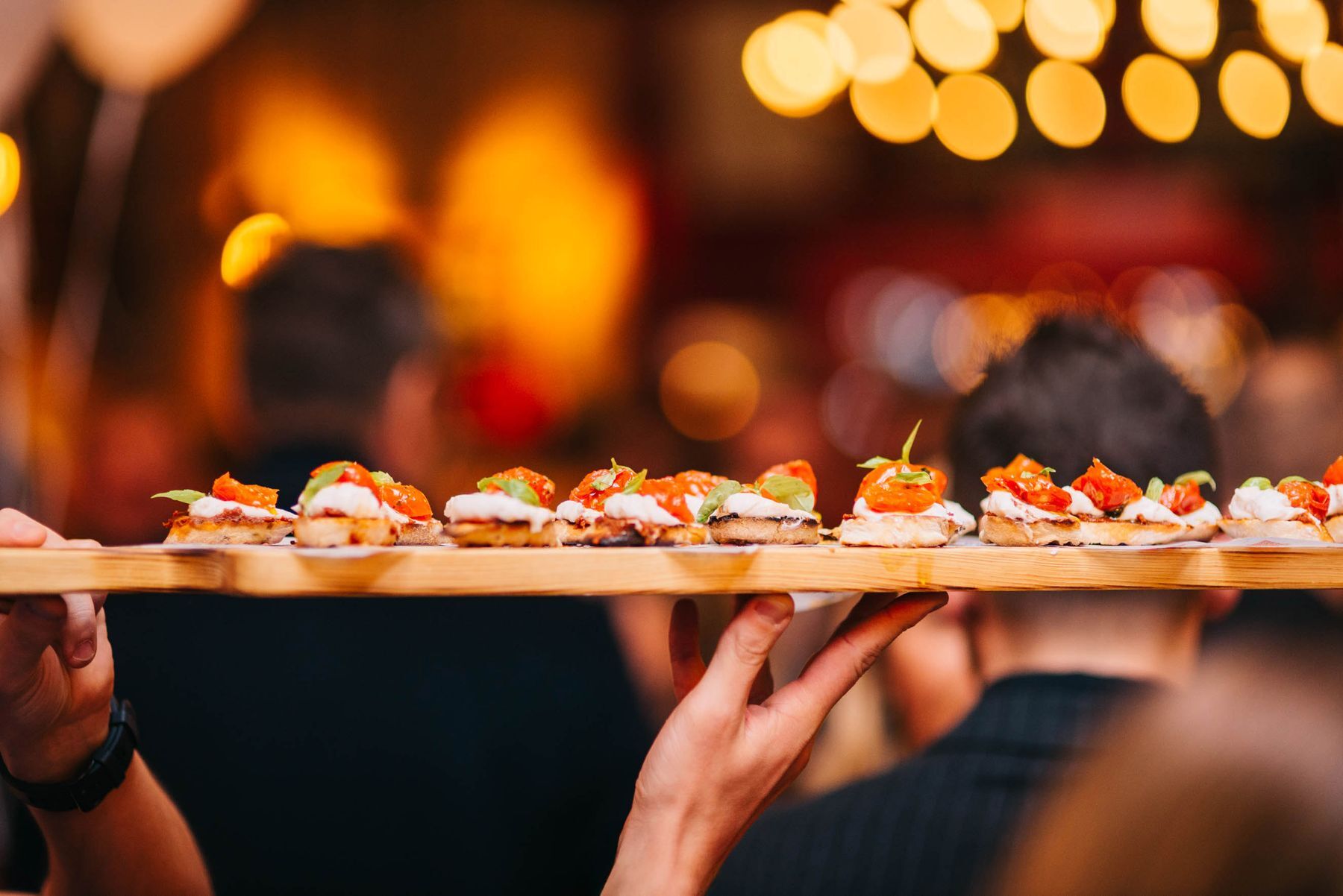 A wooden board of small bites carried through a warm, busy room